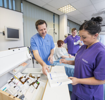 Nursing student standing in front of a medicine dispensing cabinet being shown a book by a tutor