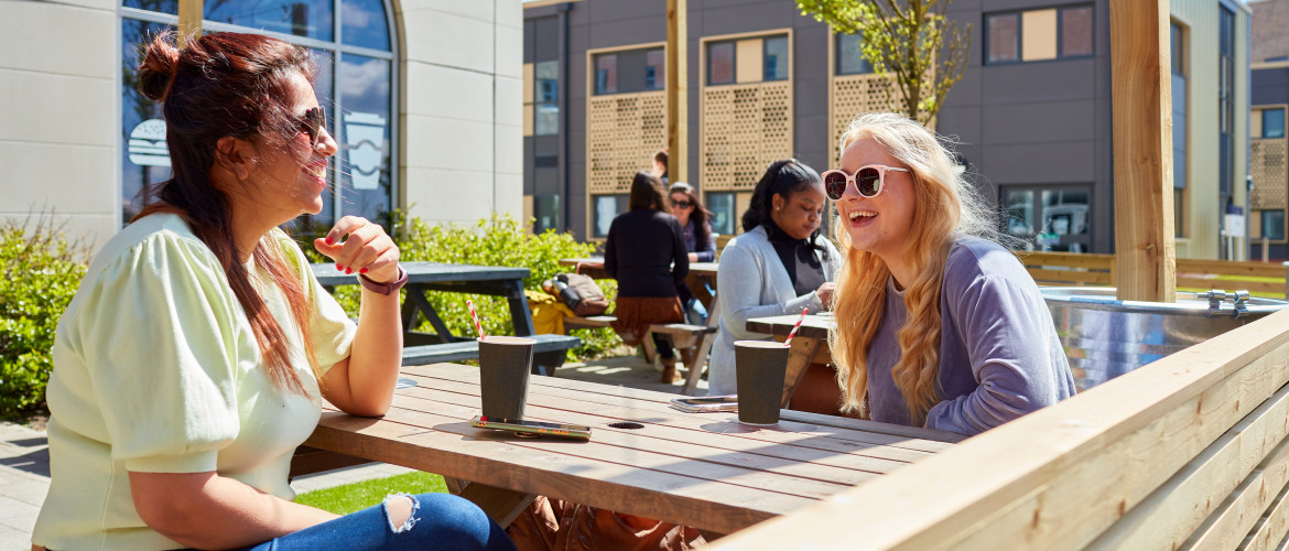 Two students sat outside on a picnic bench drinking coffee